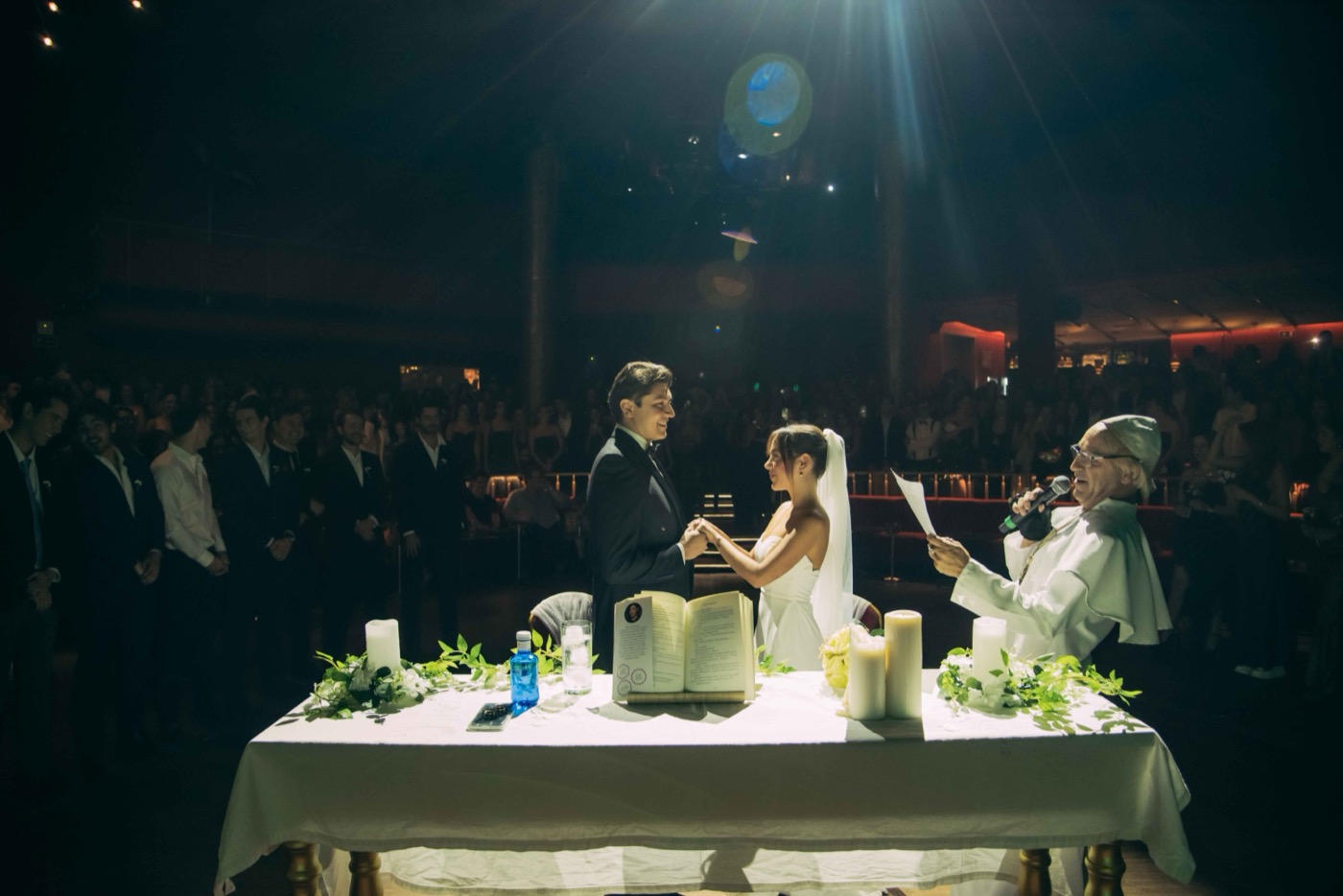 The bride and groom hold hands at the altar as the officiant reads, the whole room watching